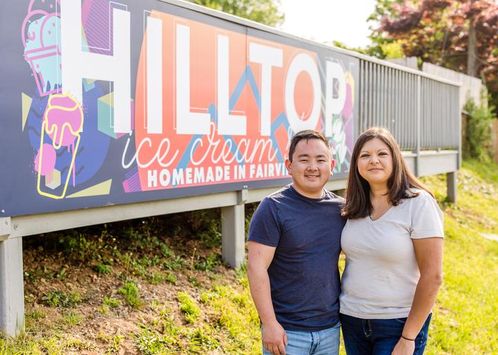 Hilltop Ice Cream Shop owners standing beside the colorful roadside shop sign